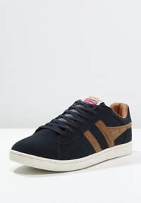 Sneaker in suede blu navy con dettagli in pelle marrone, dotato di due strisce laterali, lacci piatti e una linguetta testurizzata con logo e Union Jack.