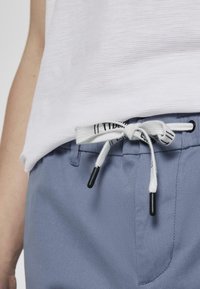 Pantalones de color azul claro con un acabado texturizado, que presentan un cordón blanco con acentos negros y una parte superior blanca con costillas parcialmente visible.