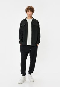 Sort skjorte med korte ærmer og to brystlommer, parret med sorte sweatpants og hvide sneakers. Stoffet ser struktureret og afslappet ud.