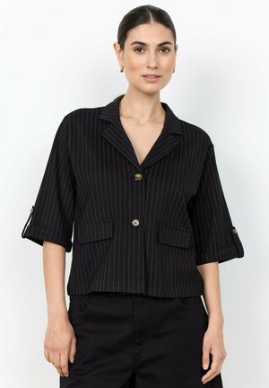 Vrouw met donker haar naar achteren gebonden draagt een zwart krijtstreep kort mouwen blazer met gouden knopen en bijpassende zwarte broek tegen een eenvoudige achtergrond.