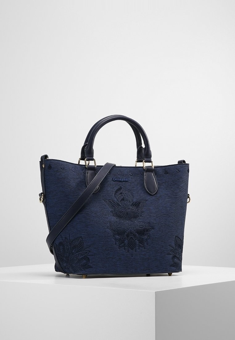 Sac à main bleu foncé avec deux poignées et une bandoulière, présentant une broderie florale et de petits clous métalliques sur une surface blanche.