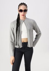 Cardigã cinza tricotado com gola redonda, fecho de botão e mangas longas. Usado sobre uma blusa branca e combinado com jeans escuros.