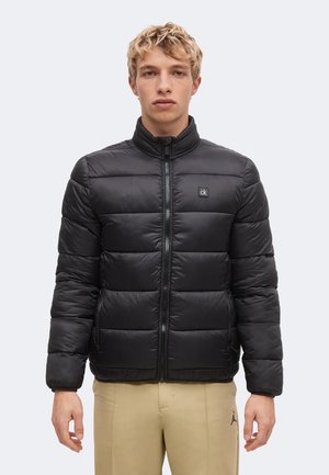 Ung mand med kort blondt hår iført en sort dunjakke med lynlås og beige bukser mod en ensfarvet lys baggrund.