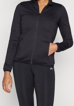 Person wearing black zip-up athletic jacket with zippered side pockets and black leggings with small logo near hip.