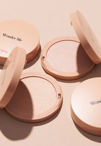 Trois boîtiers ronds de couleur pêche, étiquetés "Wonder Me" sur le dessus, contenant une poudre scintillante à la texture lisse et réfléchissante.