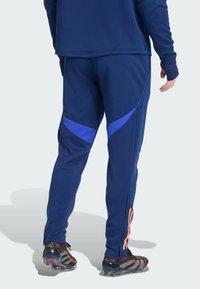 Marineblaue Sport-Trackpants mit blauen Akzenten, schmal zulaufenden Beinen und seitlichen Reißverschlüssen. Getragen mit schwarzen Fußballschuhen mit roten Details.
