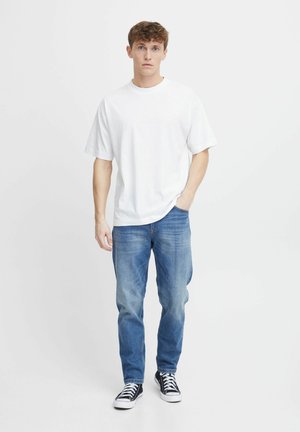 Weißes Baumwoll-T-Shirt mit kurzen Ärmeln, kombiniert mit blauen Jeans und schwarzen Sneakers. Der Stoff wirkt weich und lässig.