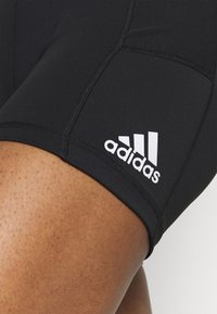 Czarne spodenki sportowe z elastycznego materiału, z białym logo Adidasa i widocznymi detalami szwów po bokach.
