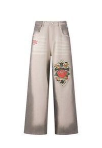 Pantalones de mezclilla de pierna ancha en beige claro con áreas desgastadas. Presenta bolsillos frontales y un parche gráfico colorido con una rosa y texto.
