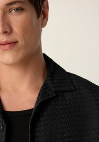 Black collared shirt with textured fabric, featuring a subtle pattern. Worn over a black crew neck t-shirt. Close-up of neckline and face.