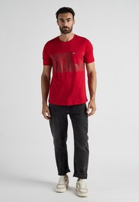 Rode katoenen T-shirt met korte mouwen, voorzien van een gestructureerd grafisch ontwerp over de borst, gecombineerd met donkere jeans en witte sneakers.