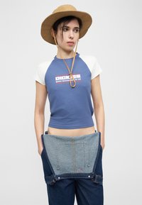 Νεαρό άτομο που φοράει καπέλο με πλατύ γείσο σε χρώμα καμηλό, μπλε και λευκό crop top Dickies, κρατώντας τζιν σαλοπέτα που έχει κατέβει στους γοφούς, μπροστά σε απλό φόντο.