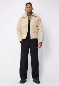 Veste en laine beige avec col, deux poches avant et fermeture éclair, associée à un pantalon noir et des chaussures. Texture lisse, design décontracté.
