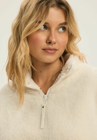 Polarowy półgolf w kremowym kolorze z zapinanym kołnierzem, charakteryzujący się teksturowaną powierzchnią oraz zamkiem błyskawicznym z logo dla dodatkowego detalu.