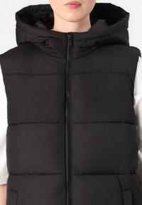 Schwarze gepolsterte Weste mit Kapuze, ausgestattet mit einem Frontreißverschluss und zwei Seiten Taschen. Glatte Textur, gestepptes Design mit horizontalen Nähten.
