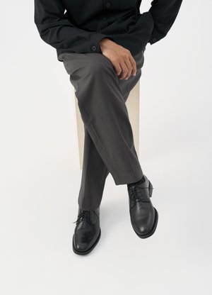 Black leather lace-up shoes with a polished finish, paired with gray tailored pants, showcased in a seated position against a white background.