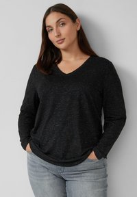 Junge Frau mit langen braunen Haaren, die einen schwarz-gemusterten V-Ausschnitt-Pullover und hellgraue Jeans trägt, steht mit den Händen in den Taschen vor einer grauen Wand.