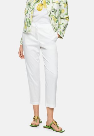 Frau trägt weiße kurze Hosen, eine zitronenbedruckte Jacke und ein T-Shirt, mit grünen und goldenen offenen Sandalen, stehend vor weißem Hintergrund.