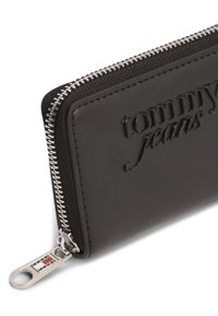 Μαύρο δερμάτινο πορτοφόλι με ανάγλυφο λογότυπο "tommy jeans", ασημένιο φερμουάρ και μεταλλική λεπτομέρεια με σχέδιο σε κόκκινο, άσπρο και μπλε.