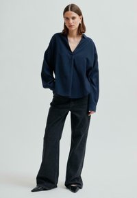 Tummansininen napillinen pusero, jossa on kaulus, yhdistettynä korkeavyötäröisiin, leveälahkeisiin mustiin farkkuihin ja teräväkärkisiin mustiin kenkiin. Suuri ja pehmeä kangas, rento istuvuus.