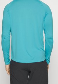 Türkises langärmliges Shirt aus glattem, dehnbarem Stoff mit Rundhalsausschnitt und engen Ärmeln, getragen über dunklen Hosen.