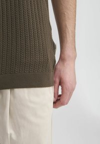 Brązowy sweter z prążkowanej dzianiny o teksturowanym wykończeniu, z boczną metką, w zestawie z jasnobeżowymi spodniami. Zbliżenie na rękę i materiał.