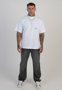 Tricou alb cu mânecă scurtă și logo brodat, asociat cu blugi drepți de culoare gri închis și adidași albi. Textură de țesătură netedă.