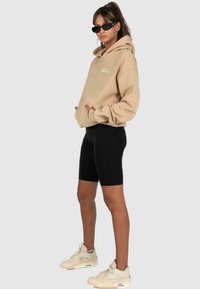 Beige mikina s kapucí a přední kapsou, doplněná černými cyklistickými šortkami a krémovými teniskami. Model nosí černé sluneční brýle a stojí na šedém pozadí.