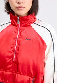 Raudona satino striukė su aukštu apykakle, pusiau užtrauktuku priekyje, kontrastuojančiomis baltomis rankovėmis, juodais akcentais ir mažuoju juodu Nike logo ant krūtinės.