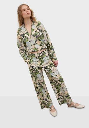 Vrouw draagt een bijpassende lange mouwen top en broek met wijde pijpen in groene, crèmekleurige en blauwe tinten, gecombineerd met crèmekleurige platte schoenen.