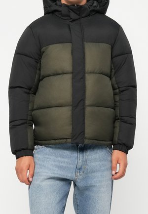 Homme portant une veste matelassée bicolore noire et vert olive avec capuche et un jean bleu, debout devant un fond uni.