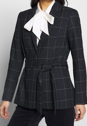 Kvinde iført en mørk ternet blazer med bælte og en hvid bluse med en stor sløjfe ved kraven.