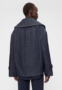 Sötét farmerdzseki nagy gallérral, széles ujjakkal és egyenes szabással. Kiemelkedő részletek között szerepelnek a gombos mandzsetták és a látható varrások.