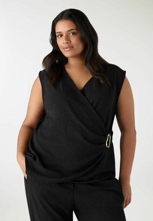 Donna con capelli scuri che indossa un top nero senza maniche con fibbia dorata e pantaloni neri, in piedi con le mani nelle tasche su sfondo bianco.