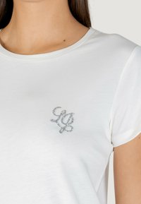 Biała koszulka z krótkim rękawem i okrągłym dekoltem; z lewej strony na piersi ozdobiona srebrnym logo z cyrkoni. Miękka tkanina bawełniana.