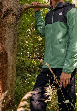Osoba ubrana w zielono-czarną kurtkę The North Face i czarne spodnie stoi w lesie, trzymając jedną ręką gałąź drzewa.