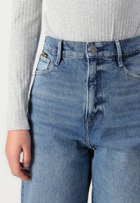 Jeans en denim bleu taille haute avec une texture décolorée, dotés d'une fermeture à bouton à l'avant, de poches latérales et d'une coupe décontractée.