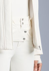 Jachetă albă, ajustată, cu o textură diagonală cordată pe mâneci, având un închidere cu nasturi tip snap și un material neted. Accentele includ accesorii din metal argintiu.