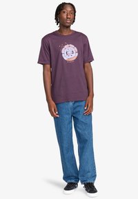 Purple cotton t-shirt featuring a graphic design and text in shades of blue and orange, paired with baggy blue denim jeans and black shoes.
