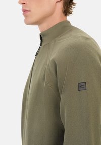 Olivengrøn zip-up jakke med h�øj krave. Ribbede tekstur og skrå syninger. Sort logo patch på venstre ærme.