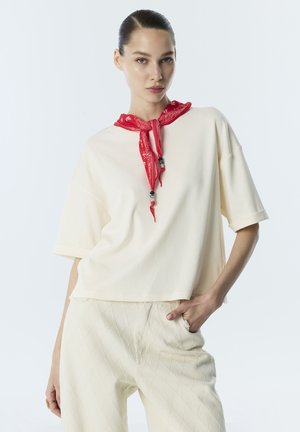 Donna con capelli pettinati all'indietro che indossa una maglia e pantaloni crema larghi, accessoriata con un bandana rossa legata intorno al collo.