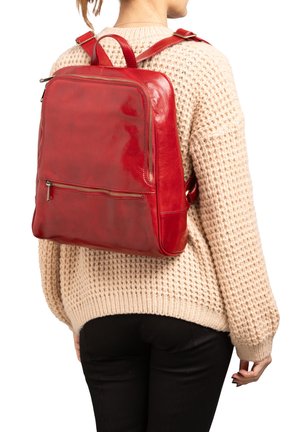 Vrouw draagt een beige trui met structuur en zwarte broek, met een grote rode leren rugzak voorzien van een voorvak met rits.