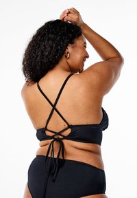 Curly-haired woman smiling with arms raised, wearing a black crisscross back bikini top and matching bottoms on a white background.