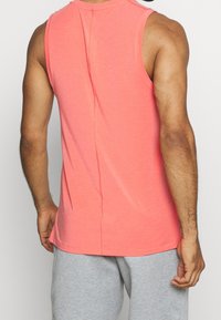 Koraloowa koszulka bez rękawów z miękkiego materiału, z tylnym szwem, noszona z szarymi dresami.