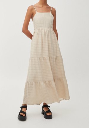 Femme portant une robe maxi beige à volants sans manches avec des bretelles spaghetti et des sandales noires épaisses avec des brides à la cheville.