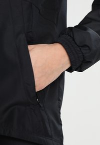 Chaqueta cortaviento negra con puños elásticos. Primer plano de una mano descansando en un bolsillo lateral, mostrando la textura de la tela y el diseño elegante.
