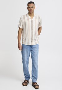 Camisa de botones de manga corta en color crema con rayas verticales marrones, combinada con jeans azul claro y sandalias marrones. Diseño sencillo, corte relajado.