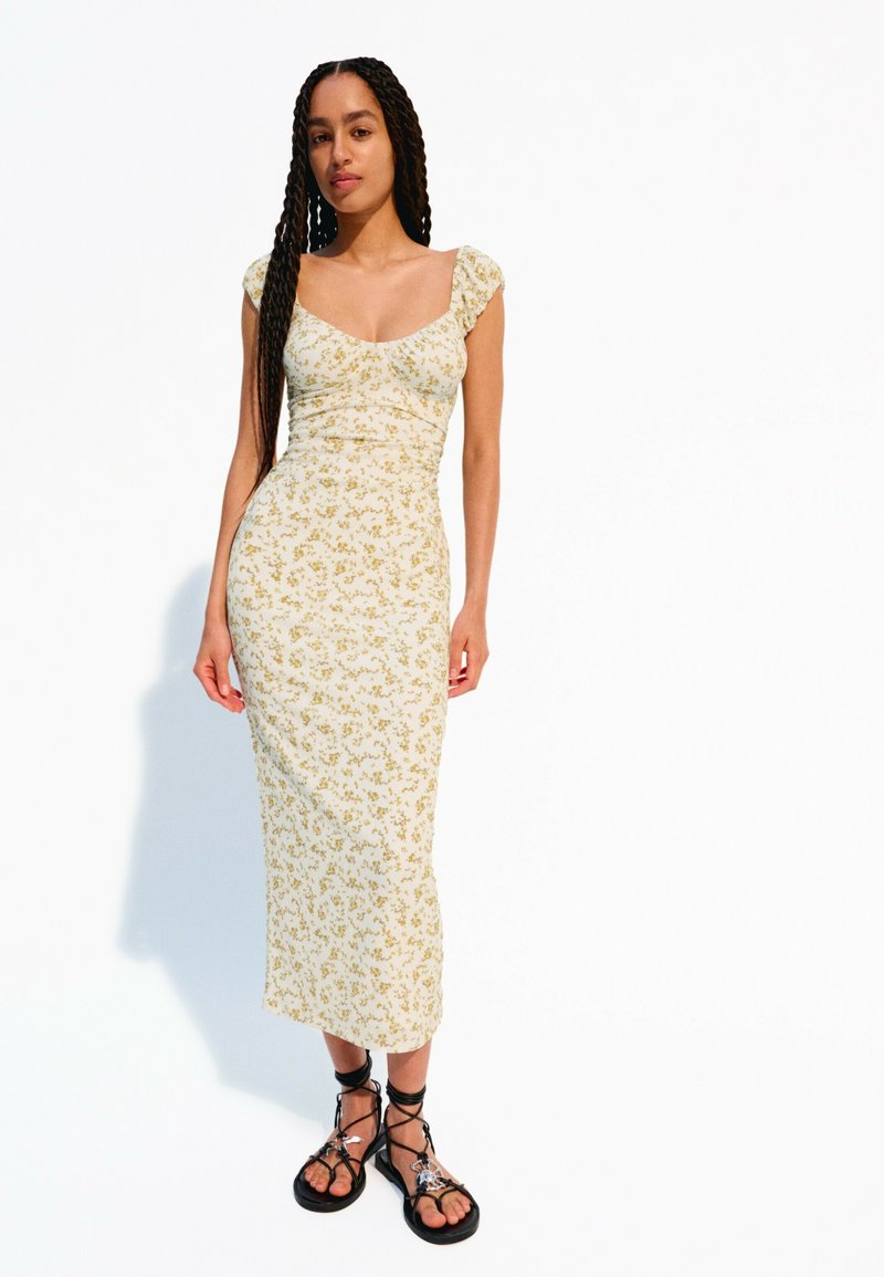 Rochie midi florală cu mâneci tip cap, de culoare crem cu motive galbene, siluetă ajustată, asociată cu sandale negre cu bretele.