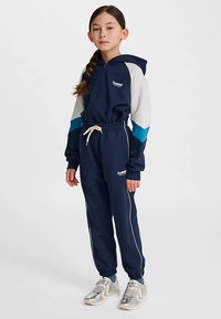 Mörkblå träningsdräkt med gråa och tealfärgade detaljer, dragkedje-hoodie, elastisk midja, sidsstreck och logotypdetalj. Silvriga sneakers.