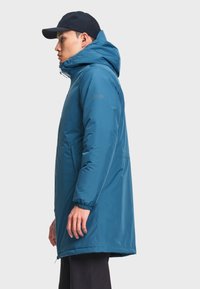 Homme portant un manteau d’hiver bleu à capuche avec des poignets élastiques et une casquette noire, debout de profil sur un fond uni.